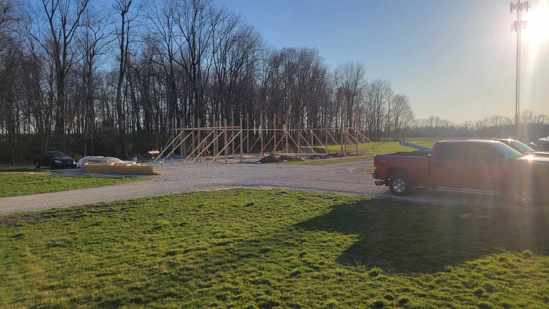 Construction site with wood framing, gravel, a red truck, and trees under a sunny sky.