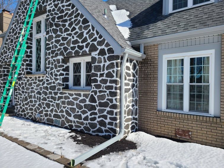 Stone and brick house with snow, gutters, and a ladder against the stone wall.