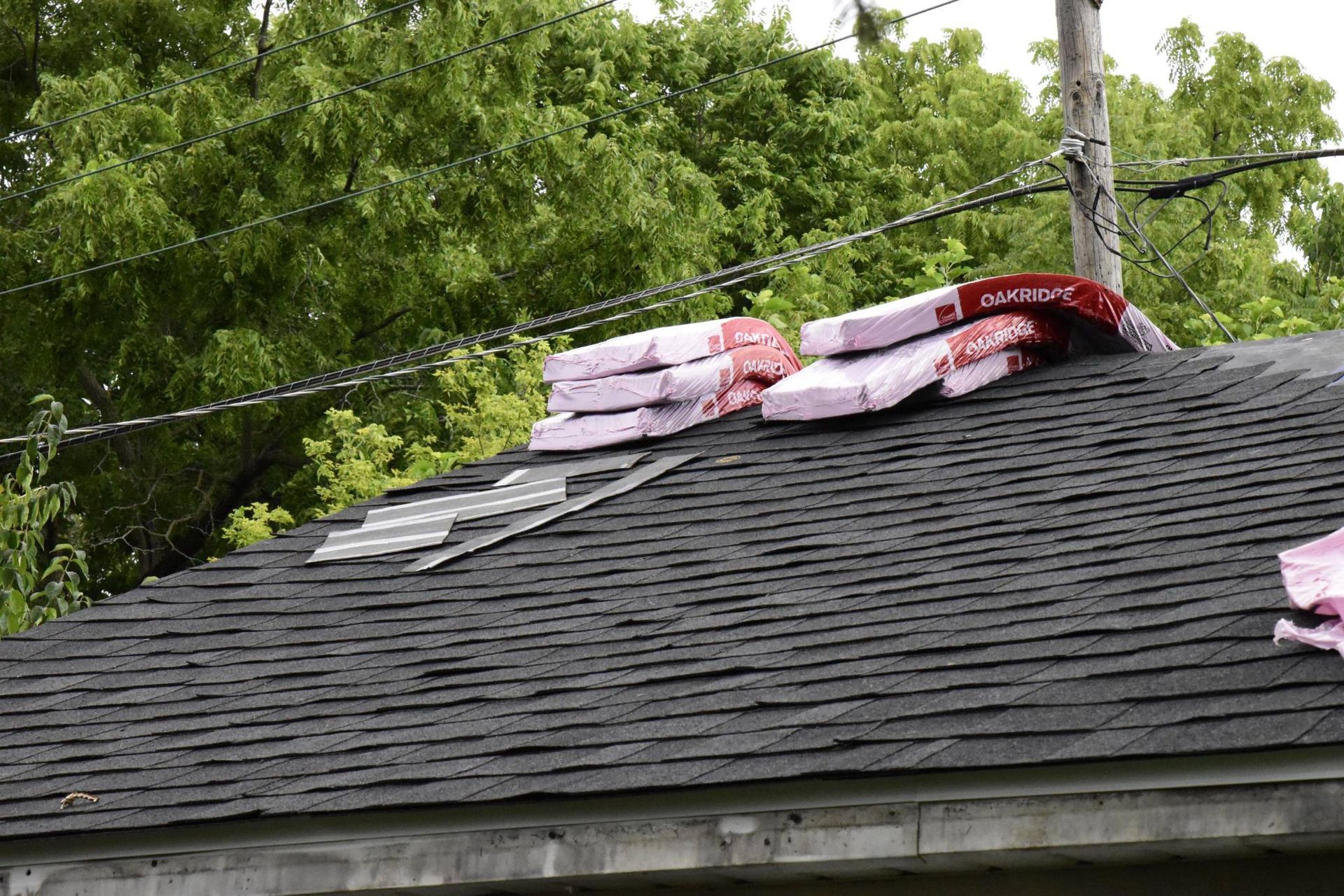 Roof with shingles, exposed underlayment, and stacked insulation against a backdrop of trees and power lines.