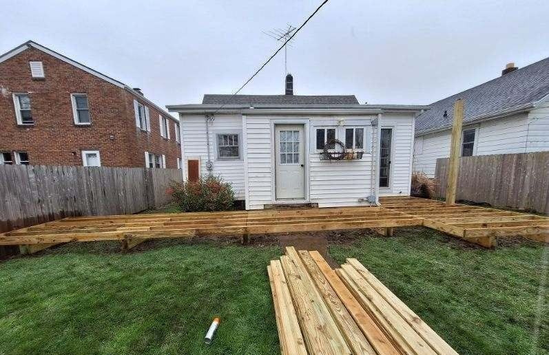 Backyard with unfinished wooden deck being constructed near a small white house and a wooden fence.