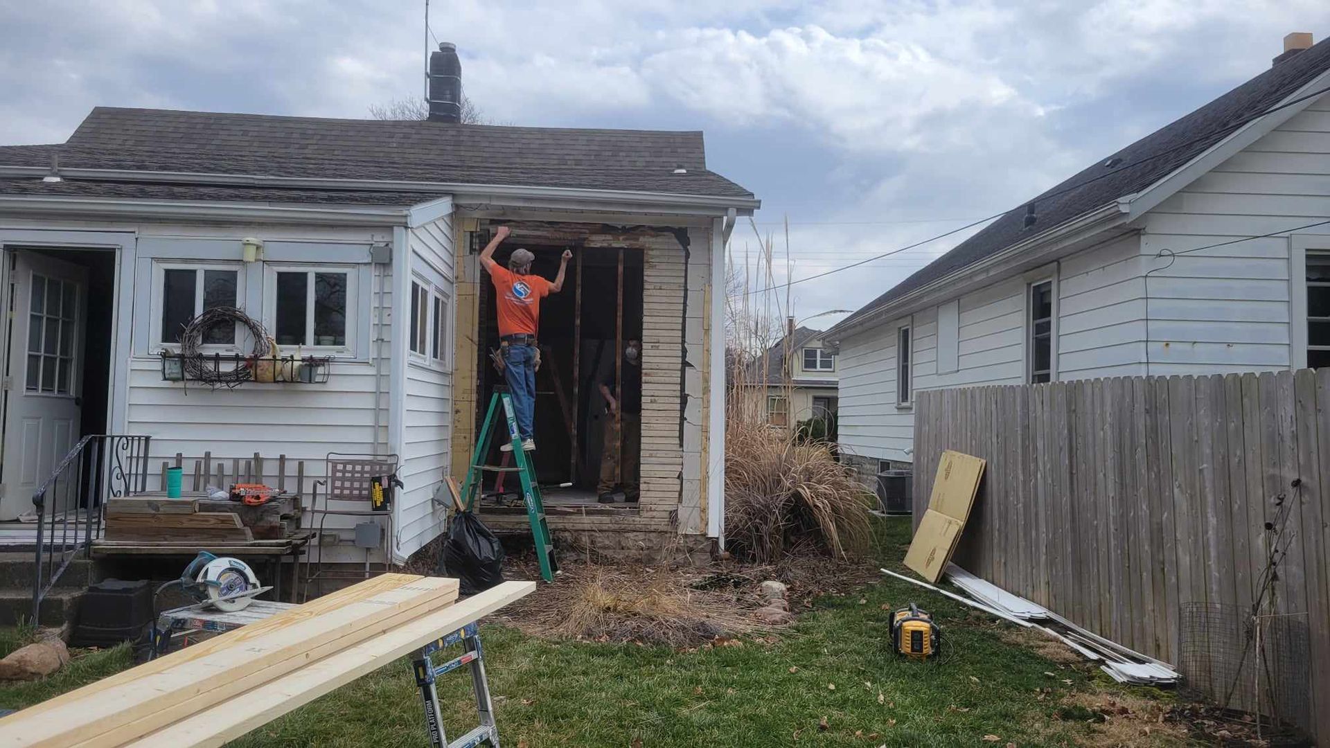 Man on ladder working on exterior of a house, with exposed frame. Cloudy day.
