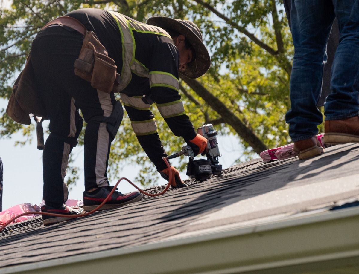 Roofer in safety gear nails shingles onto a roof; another person stands nearby.