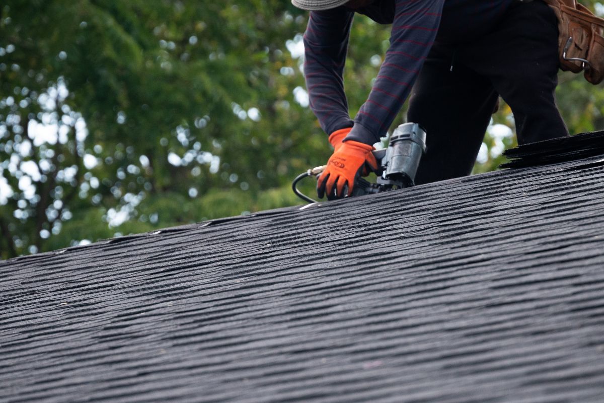 Roofer installing shingles on a roof with a nail gun, wearing orange gloves.