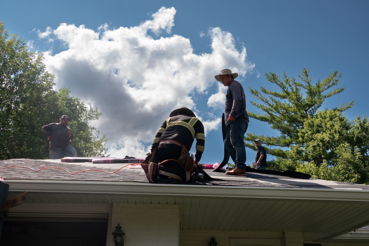 Three workers on a roof, removing shingles. Blue sky and clouds overhead.