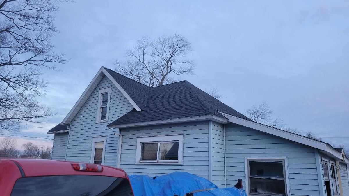 Blue house with dark roof and bare tree against a cloudy sky. A red truck is visible in the foreground.