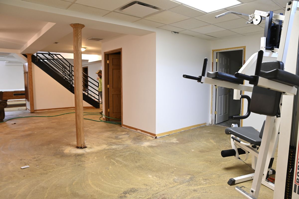 Home gym with exercise equipment, stairs, support columns, and bare concrete floor.