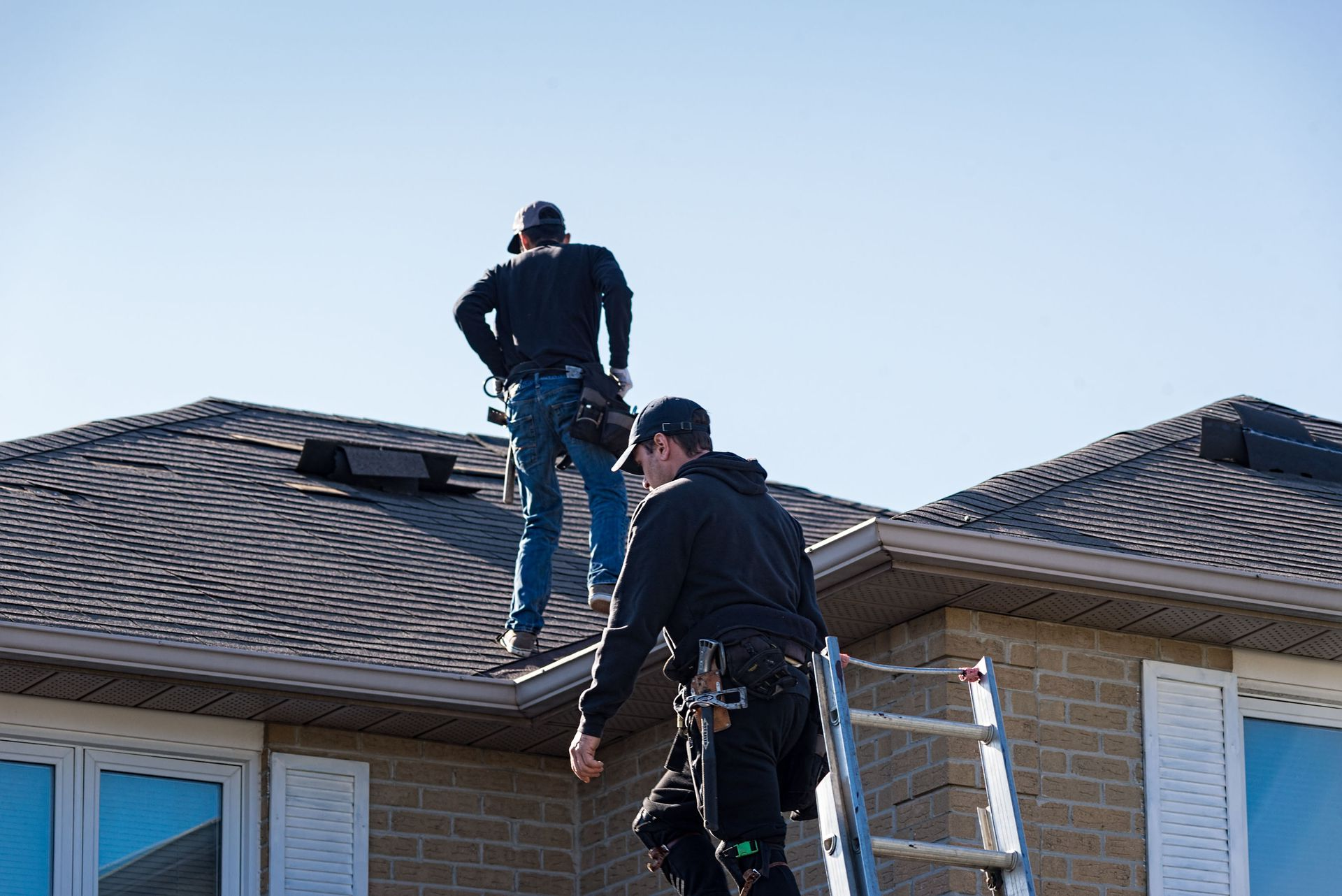 Two roofers on a roof, one descending a ladder. Blue sky, brick house, working.