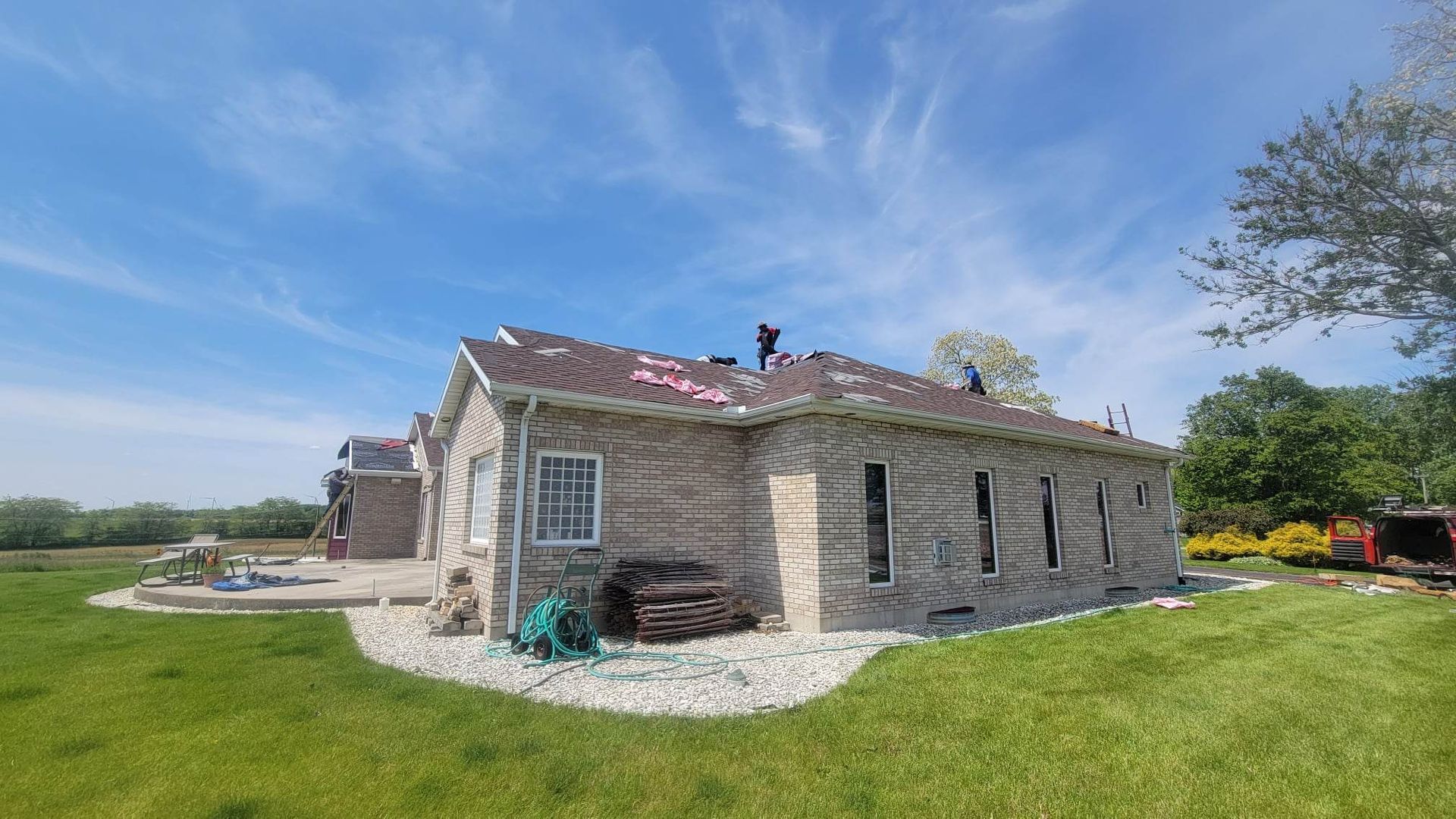 Workers on a roof of a brick house under a blue sky, some shingles removed. Green grass surrounds.