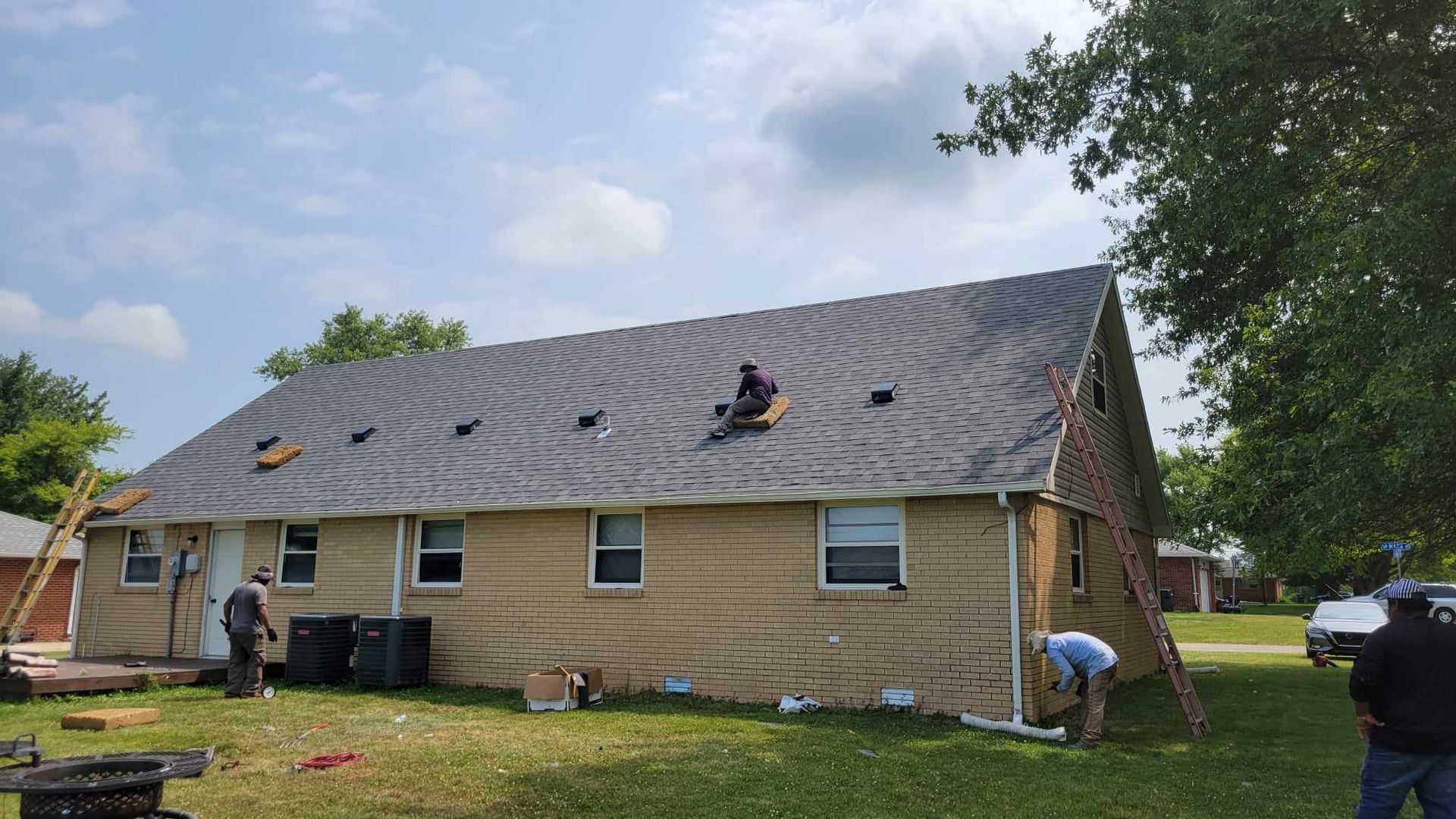Workers installing shingles on a house roof on a sunny day. Ladders, tools, and a truck are present.