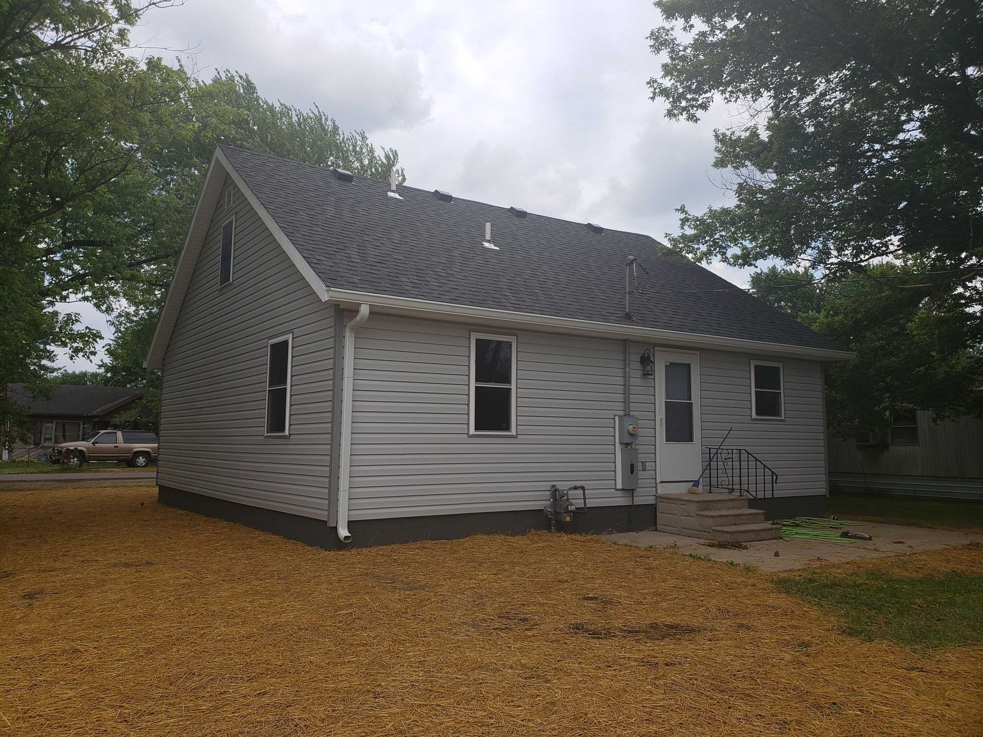 Small house with gray siding, dark roof, two windows, and door, surrounded by trees and dry ground.