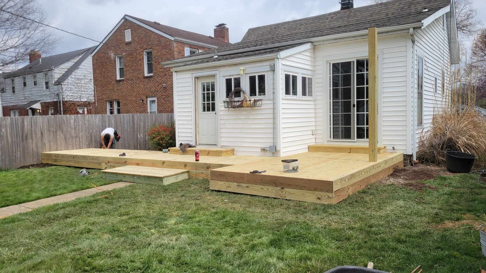 New wooden deck built on the side of a white house with a person working on it; green grass.