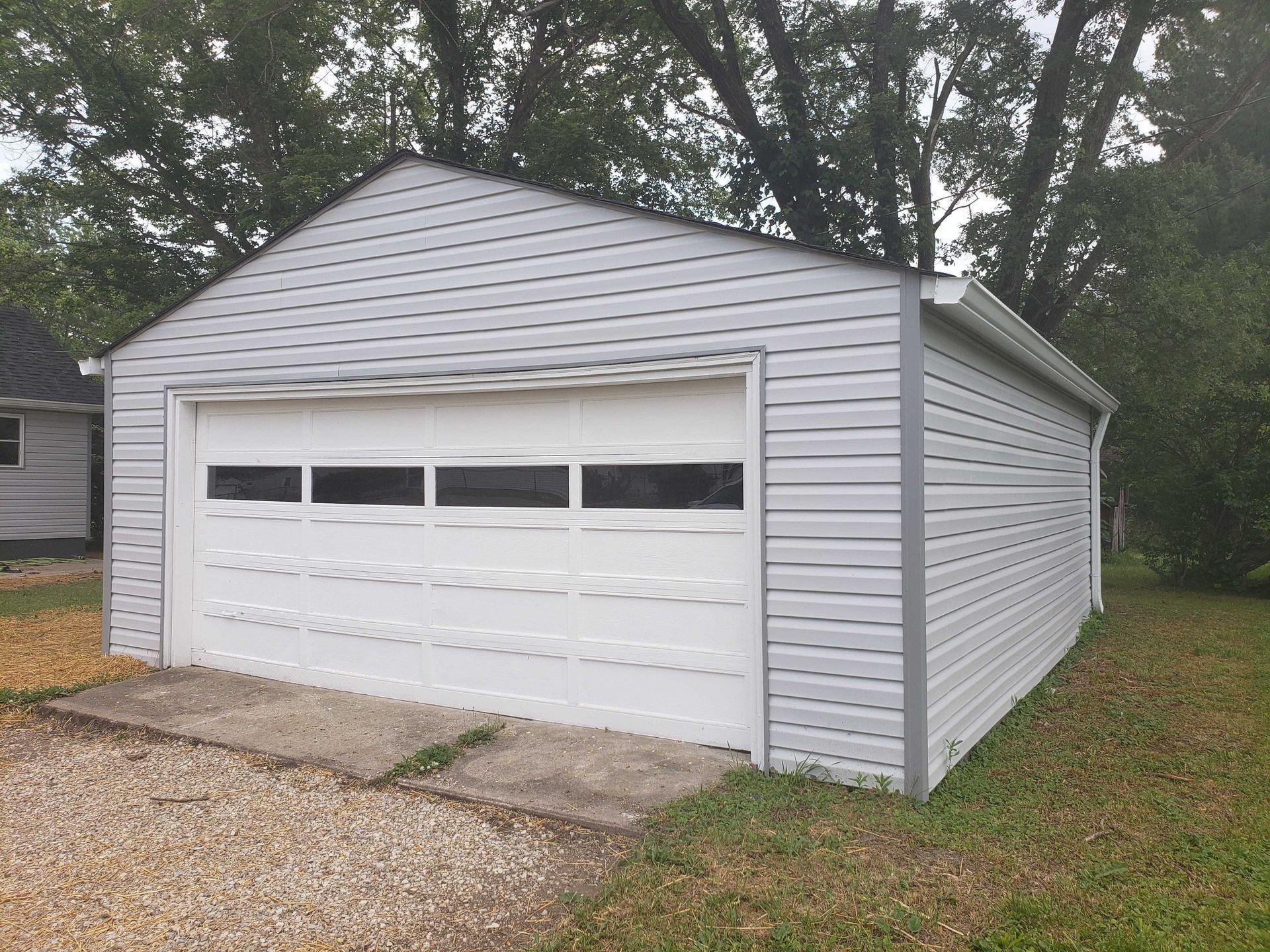 A detached garage with a white overhead door and gray siding, set on a gravel driveway and grass.