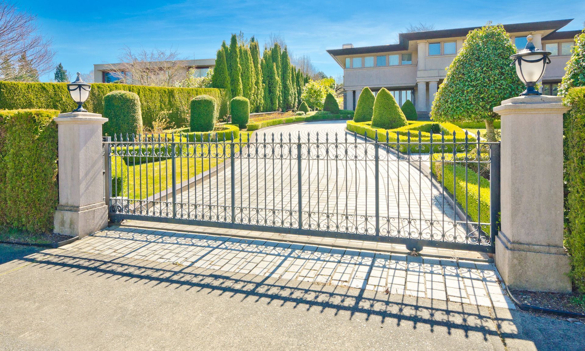 Gated driveway leading to a large suburban house with manicured hedges and bright sunlight