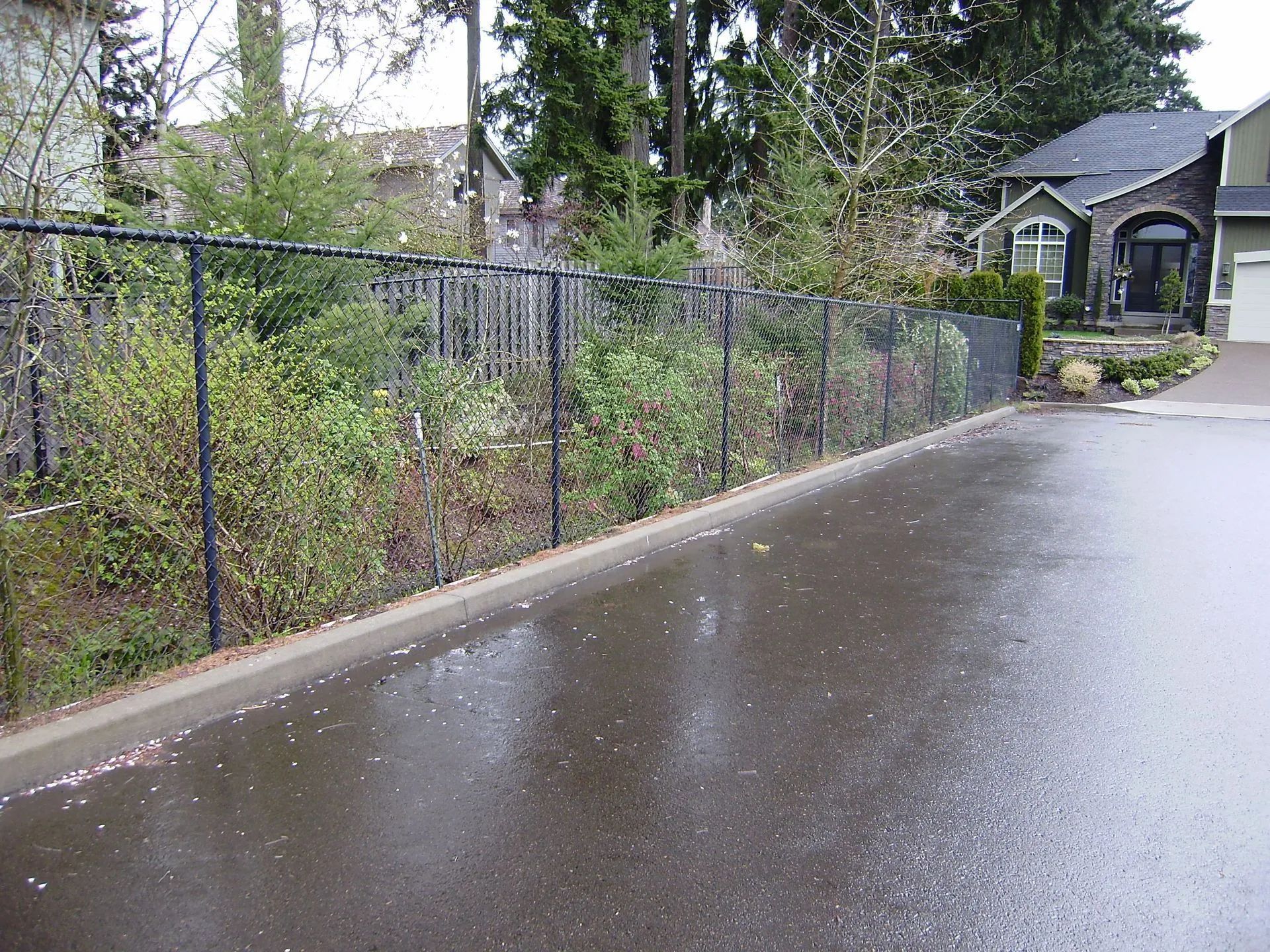 Wet driveway beside a chain-link fence, with bushes and a house in the background.