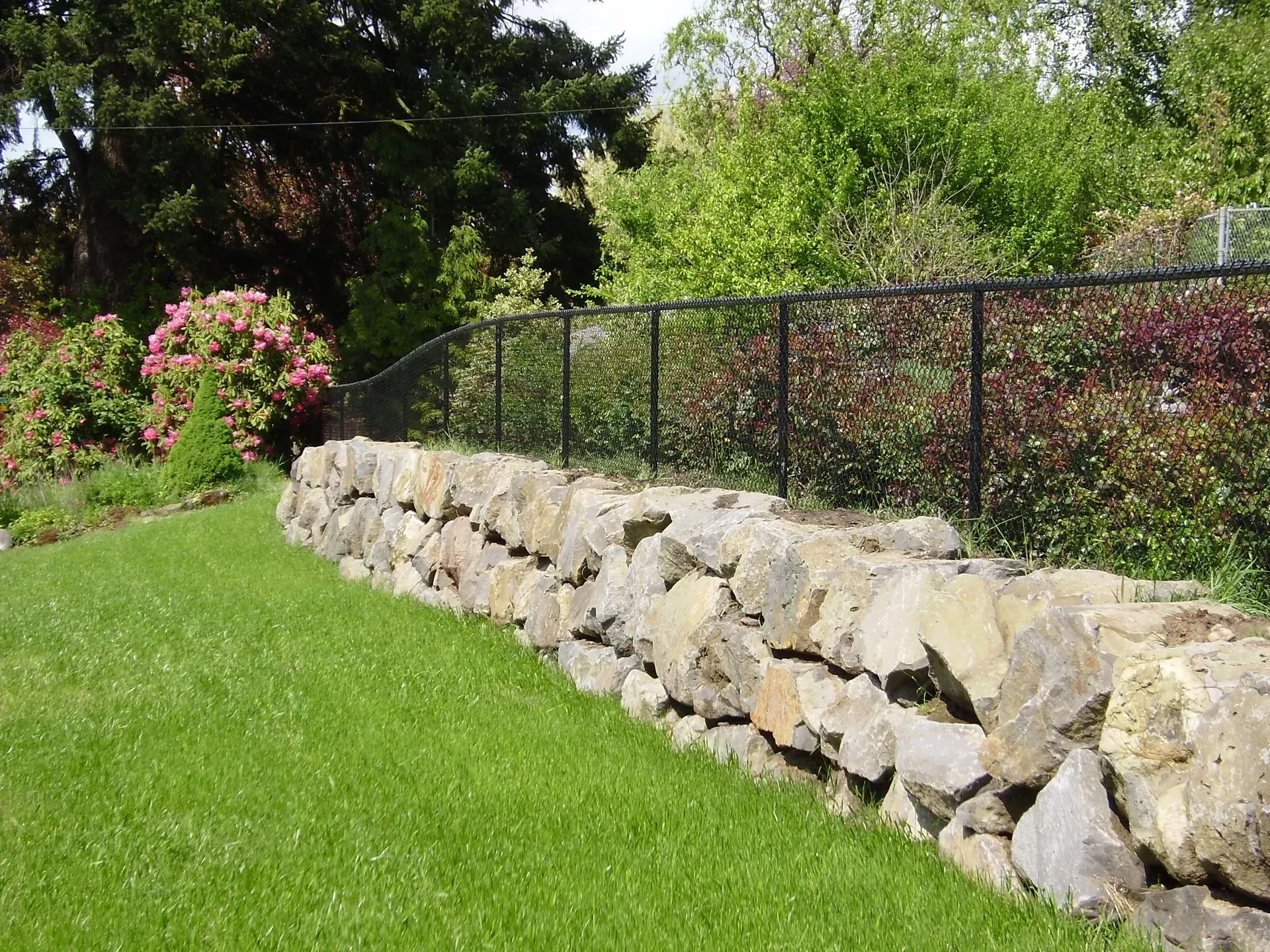 Stone retaining wall with black fence beside a green lawn and trees