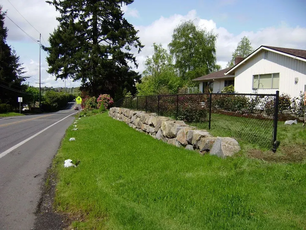Quiet suburban road beside a grassy verge, stone retaining wall, and a house with trees and fence.