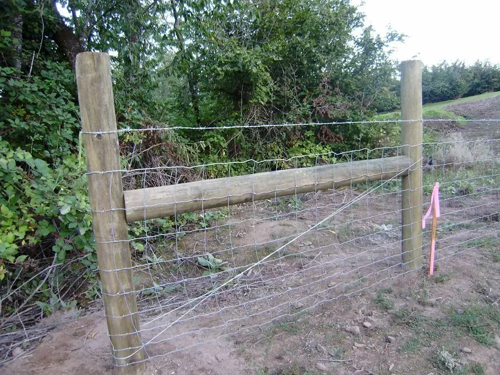 Wooden farm gate with wire fencing beside a dirt path and green shrubs.