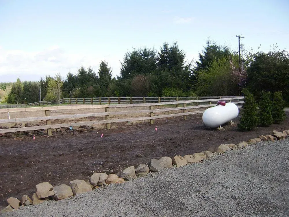 Gravel driveway beside a fenced yard with trees and a white propane tank