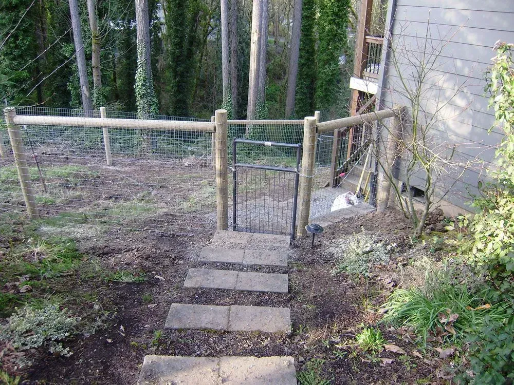 Narrow garden gate with concrete stepping stones, wooden fence, and trees along a dirt path.