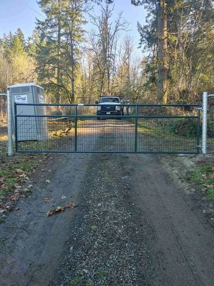 Chain-link gate across a gravel driveway with a portable toilet on the left and trees in the background