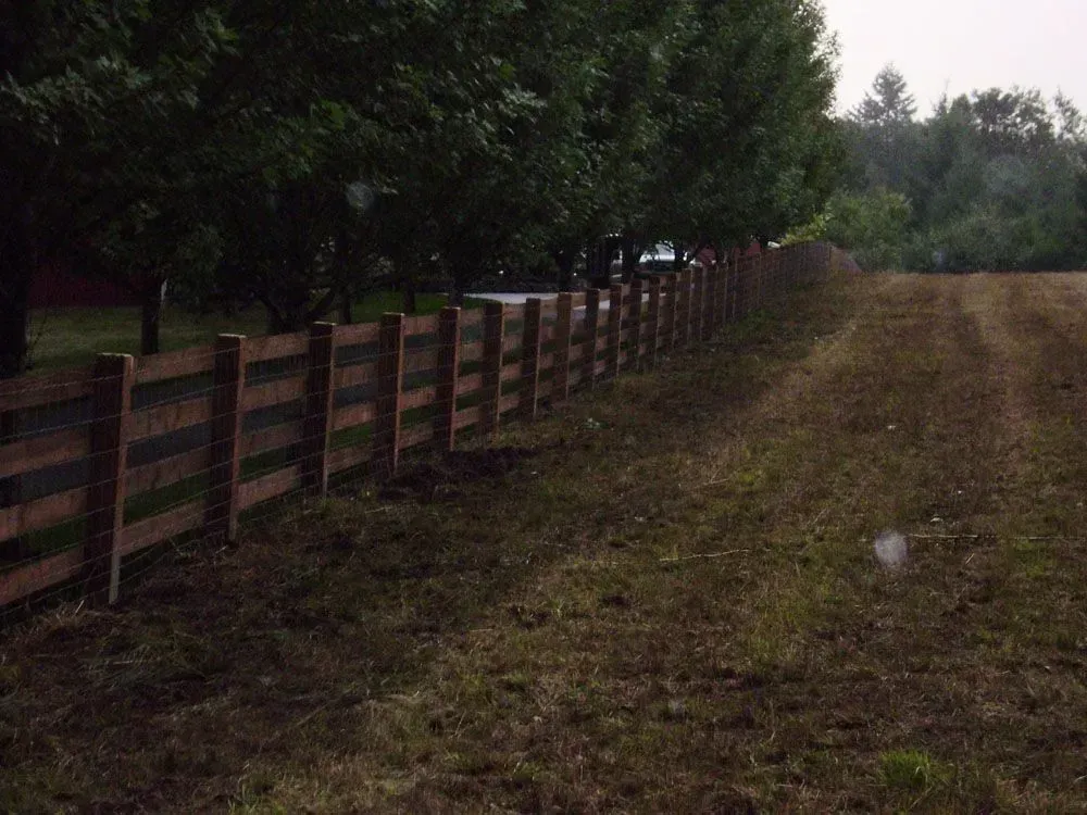 Wooden fence beside a muddy field, with trees lining the left side and overcast sky.