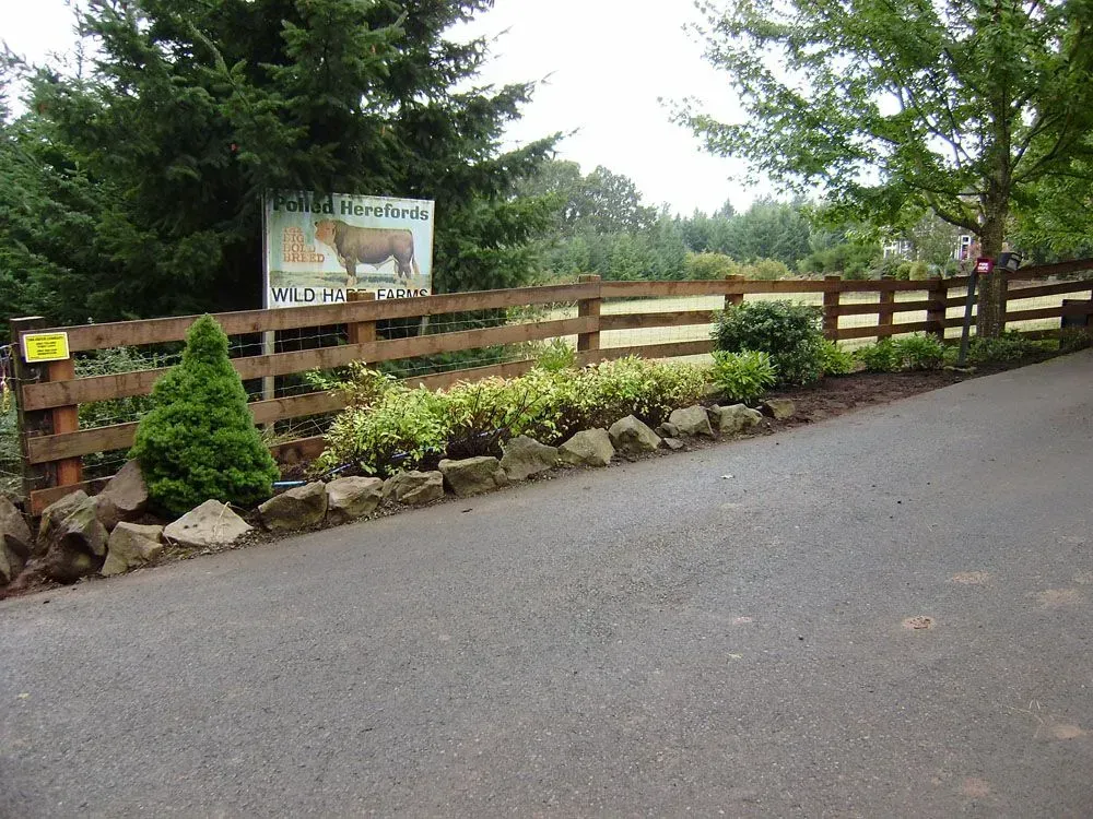 Wooden fence and stone-lined driveway entrance with a sign in a rural, tree-filled setting