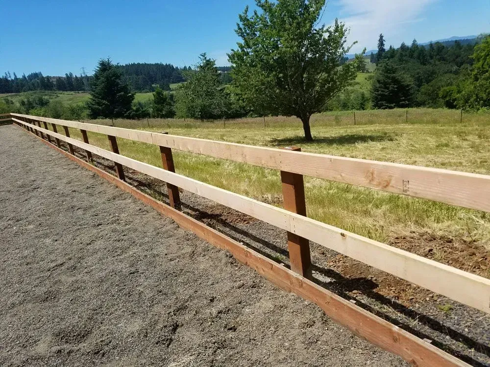 Wooden fence along a gravel path beside a grassy field with trees and hills under a clear sky