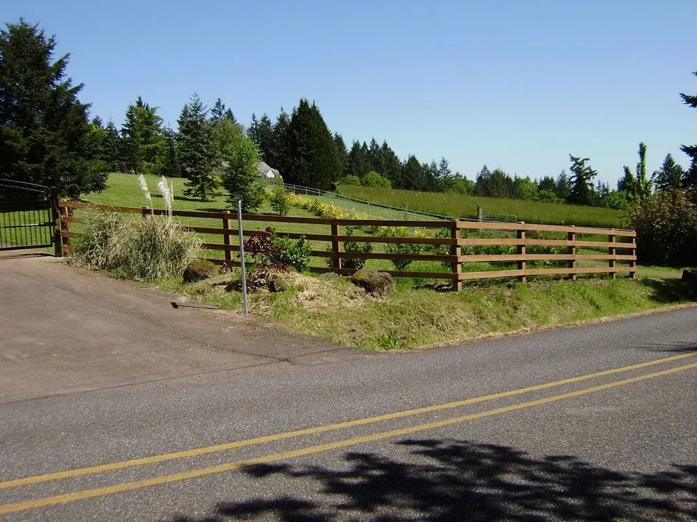 Wooden fence along a grassy roadside hill with evergreen trees under a clear blue sky