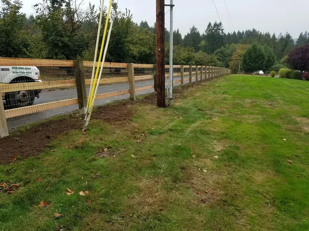 Grassy roadside beside a wooden fence, utility poles, and parked vehicles under cloudy skies.