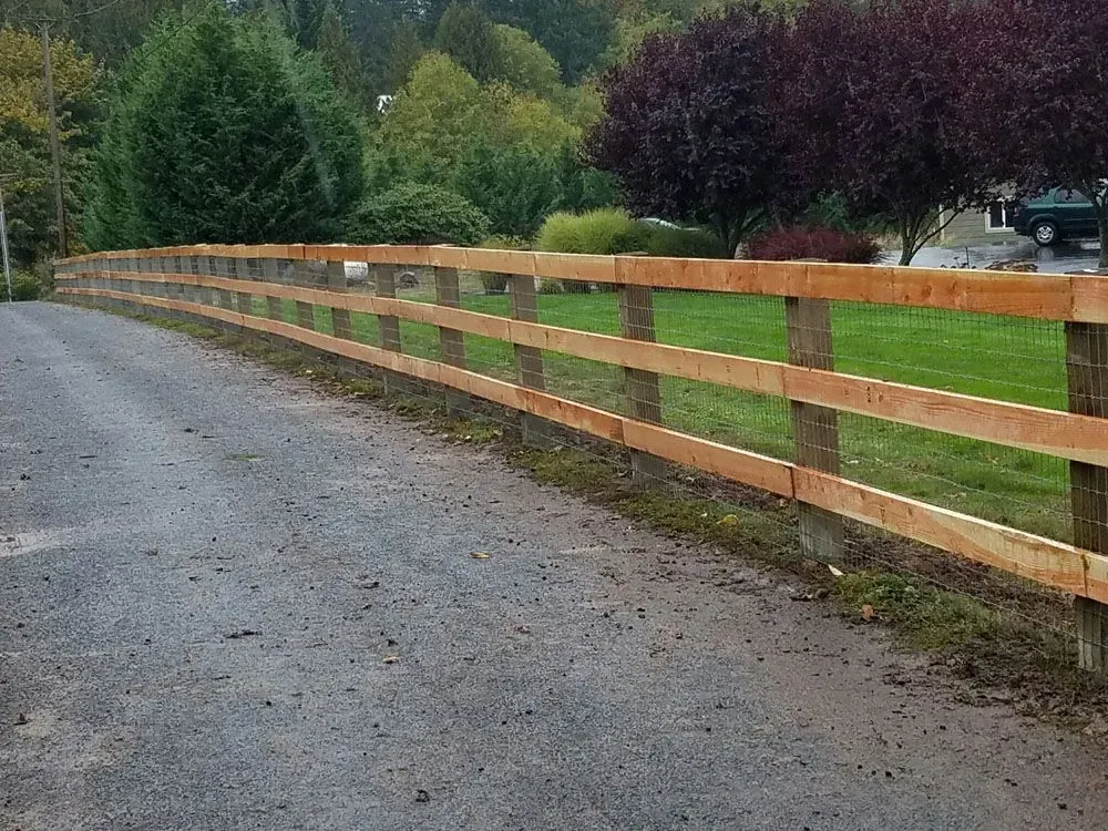 Gravel driveway beside a long wooden fence, with green fields and trees under an overcast sky.