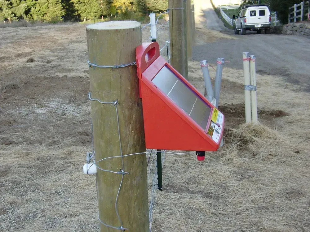 Red emergency call box mounted on a wooden post in a dry field, with fence posts and a vehicle in the background