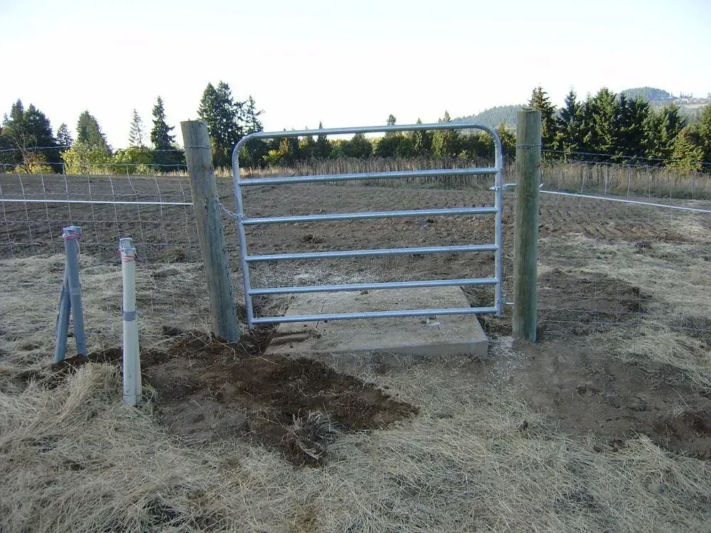 Metal gate in a dry field beside posts, with trees and hills in the background.