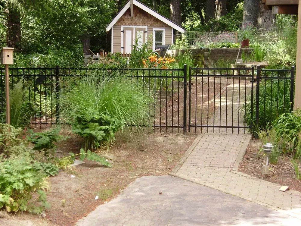 Garden path leading to a black metal gate, with a small cottage and lush greenery beyond.