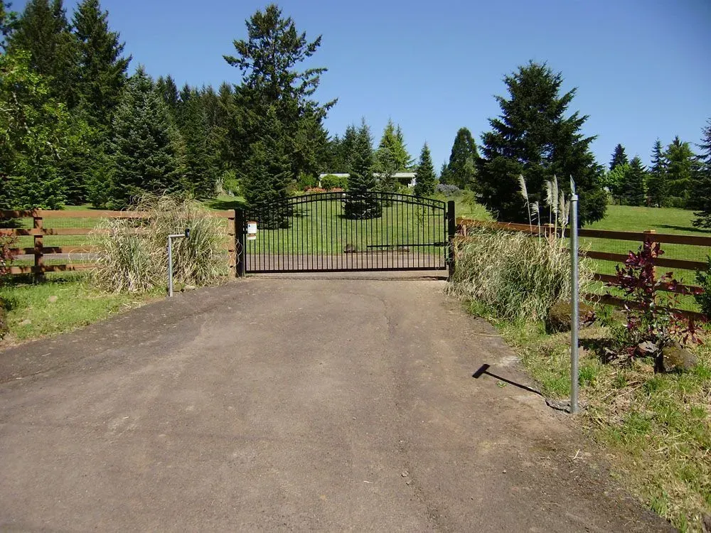 Gravel driveway leading to a closed black gate amid green trees and fencing