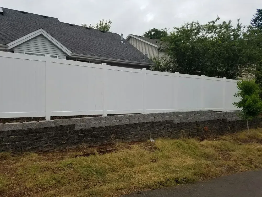 White vinyl fence atop a stone retaining wall beside a grassy roadside, with house roofs in the background