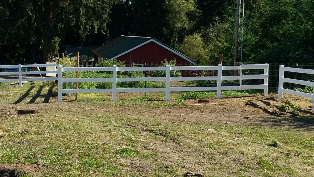 White wooden fence in front of a red barn on a grassy slope with trees in the background