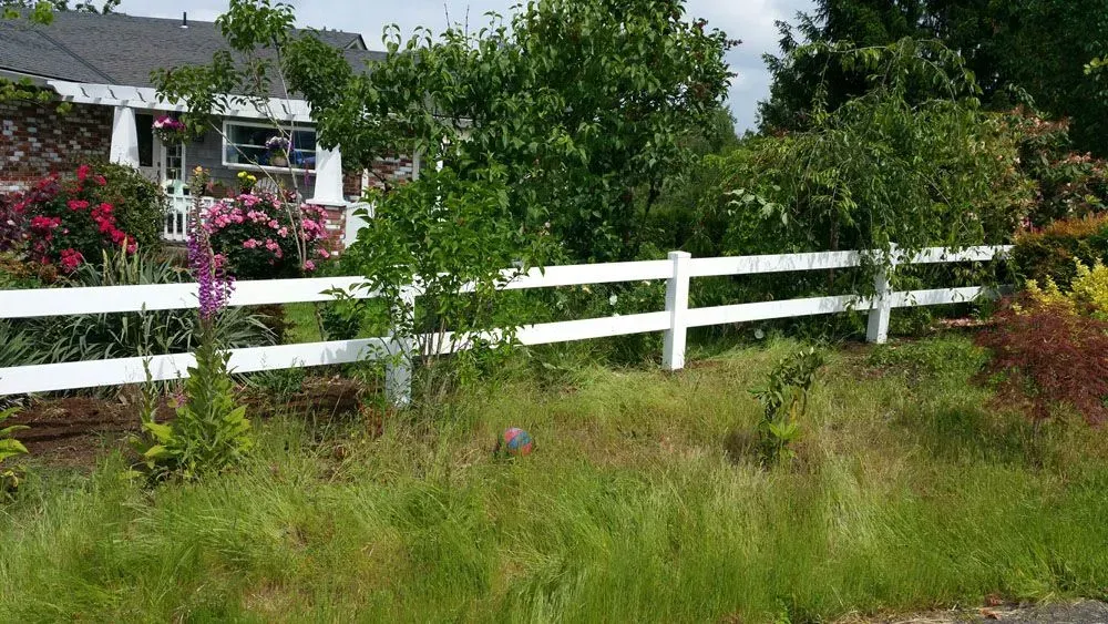 White fence beside a lush garden with pink flowers and a house in the background