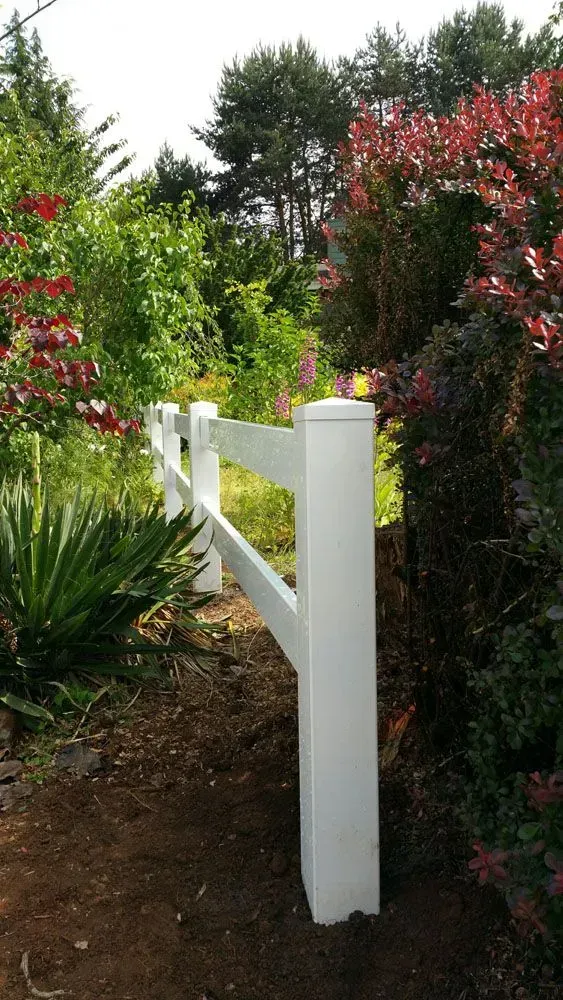 White fence post and rail beside a garden path, surrounded by green shrubs and red flowering bushes