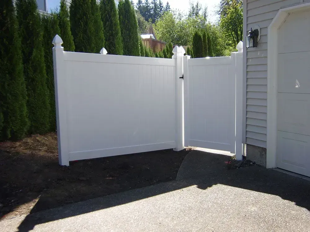 White vinyl fence and gate beside a house, opening onto a driveway with tall green hedges.