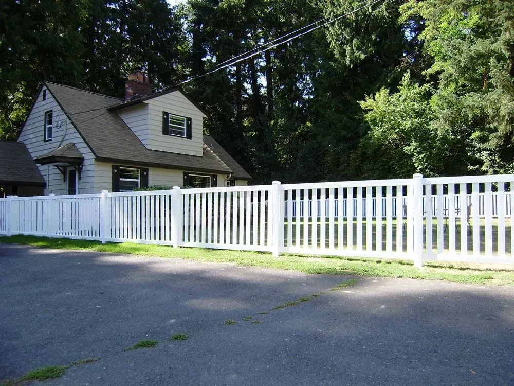 White picket fence along a driveway in front of a small house with trees in the background.