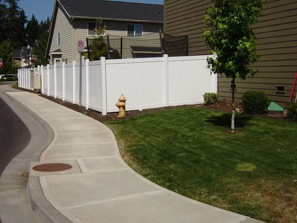 Curved suburban sidewalk beside a white fence and grassy yard with houses