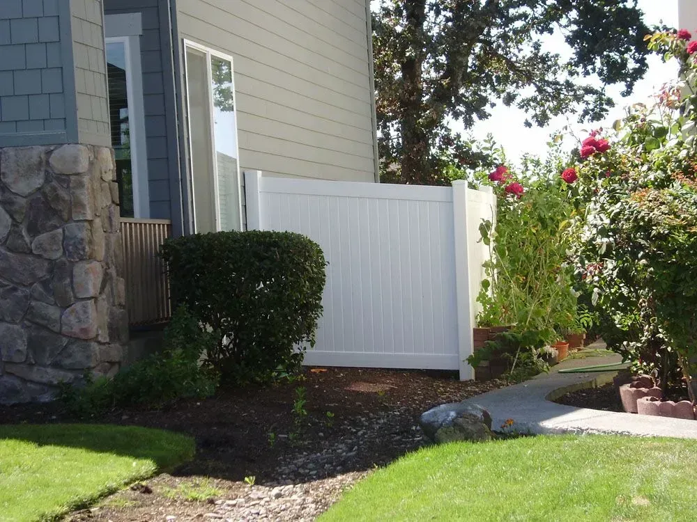 Front yard with white fence, stone house wall, shrubs, and a green lawn beside a walkway.
