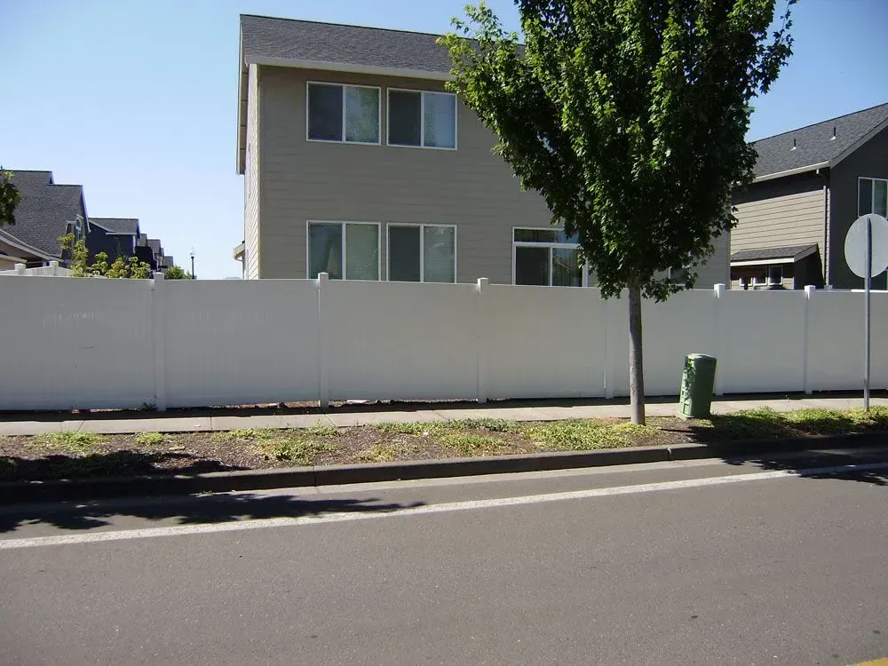 Suburban house behind a white fence, with a tree and sidewalk along a quiet street.