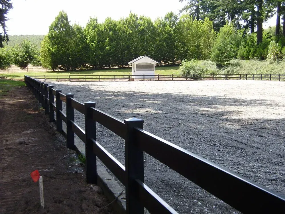 Black fence along a gravel riding arena with trees and a small white shelter in the background
