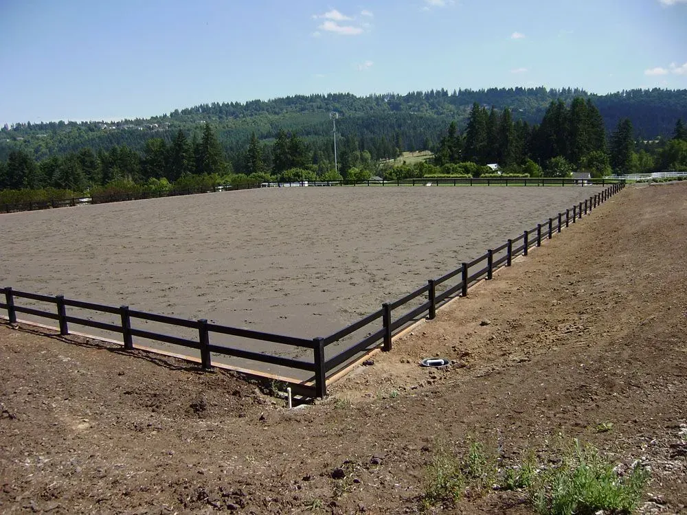 Empty fenced dirt arena with a black wooden rail fence and forested hills in the background