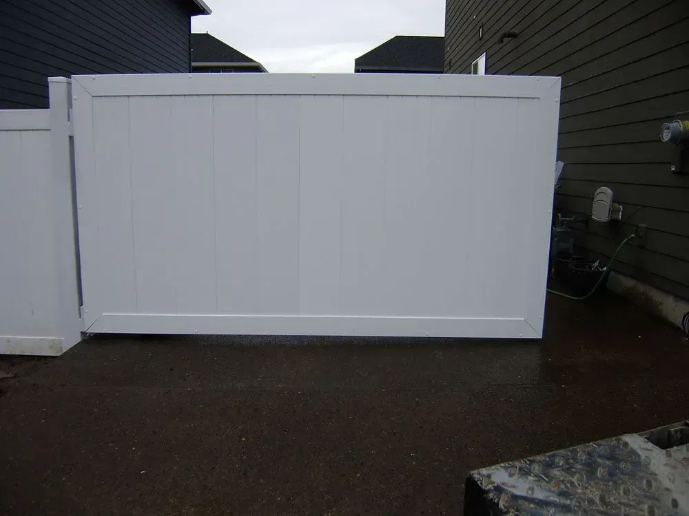 White vinyl fence panel across a wet driveway between dark-sided houses