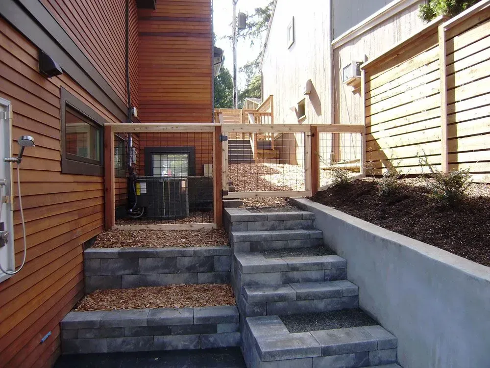 Narrow outdoor stairway between buildings with wooden siding and a raised concrete planter wall