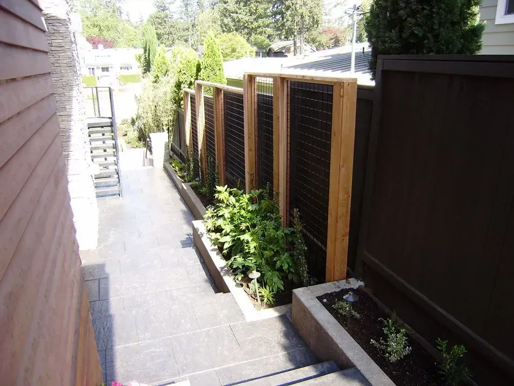 Narrow outdoor side yard with gray paving, wooden fence panels, and small garden plants along the right side.