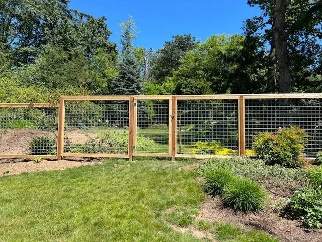 Wooden garden fence with wire mesh in a sunny landscaped yard, backed by trees and shrubs