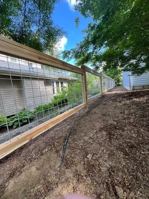 Wooden fence along a dirt path beside a house, with trees and blue sky overhead.